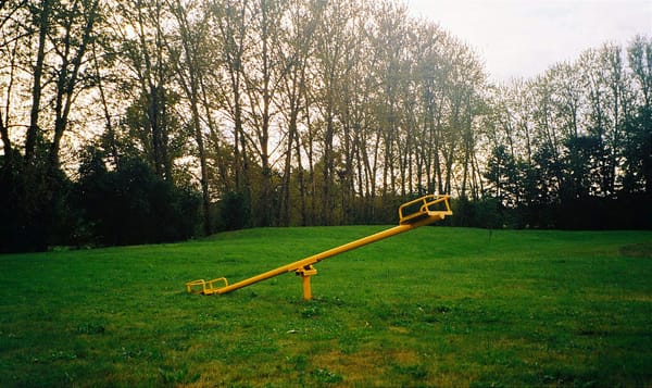 a yellow seesaw planted in the middle of a grassy field. thin trees line the periphery. their leaves and branches practically fade into the bright sky.
