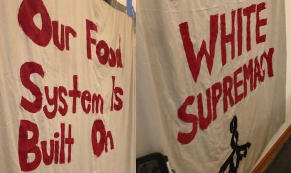 a photo of a large white sheet turned into a hand-painted banner. in red paint, the lettering reads, "Our Food System Is Buil