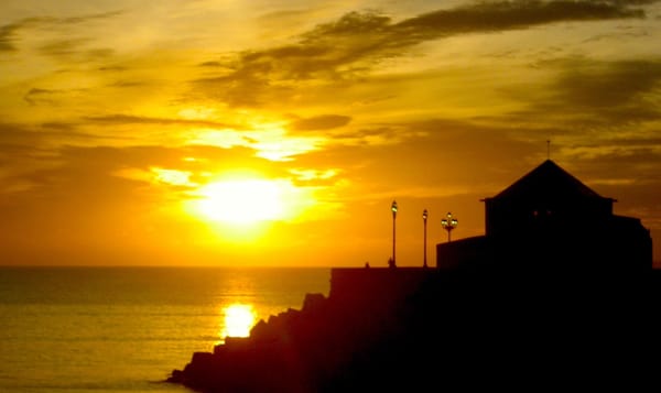 a photo of a sunset on the rocky shores of Cádiz, Spain. the still-bright yellow sun tints the sky golden. a building with a