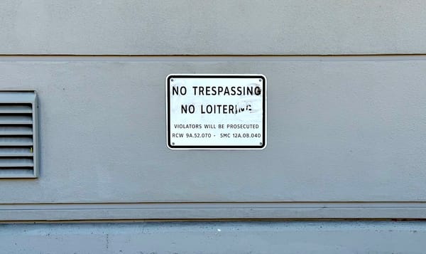 a sign against a gray building wall that reads NO TRESPASSING NO LOITERING with a threat of prosecution.