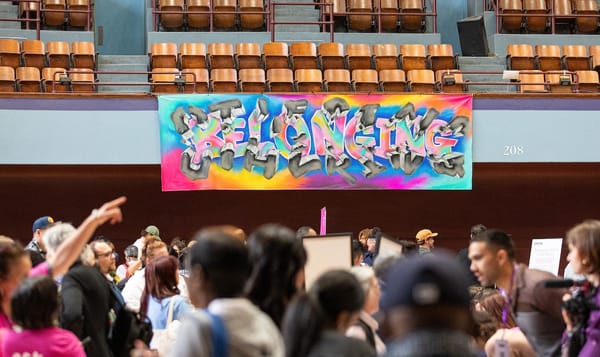 people talking together in a gymnasium with a hand painted sign above them that says "belonging" in graffiti script