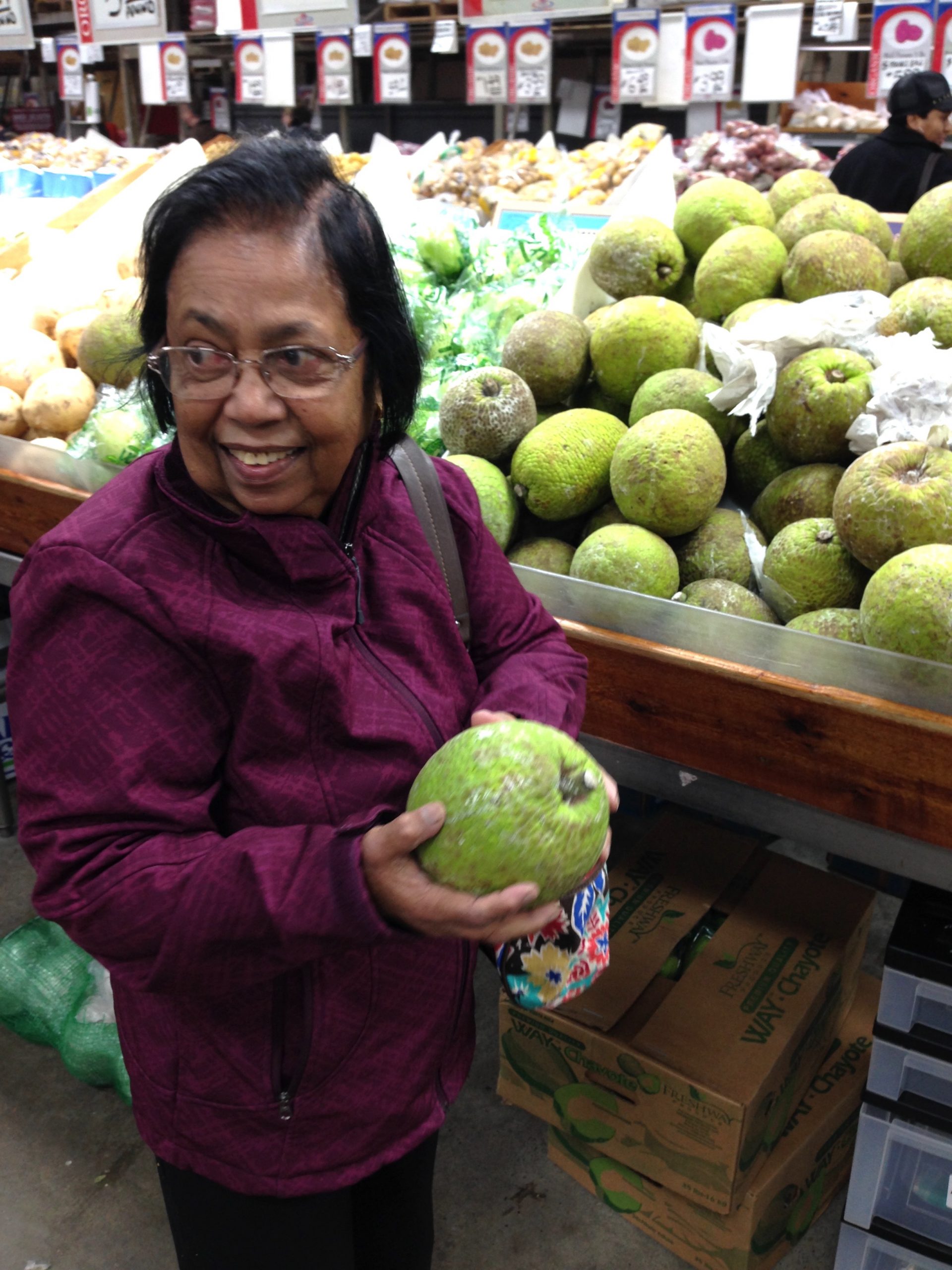 a photo of my achchi in a busy produce market, her face full of pleasant surprise. she is wearing a purple jacket and holding a breadfruit the size and shape of a green coconut. she's standing in front a whole display of them. a display that, by the look on her face, she was clearly not expecting to see.