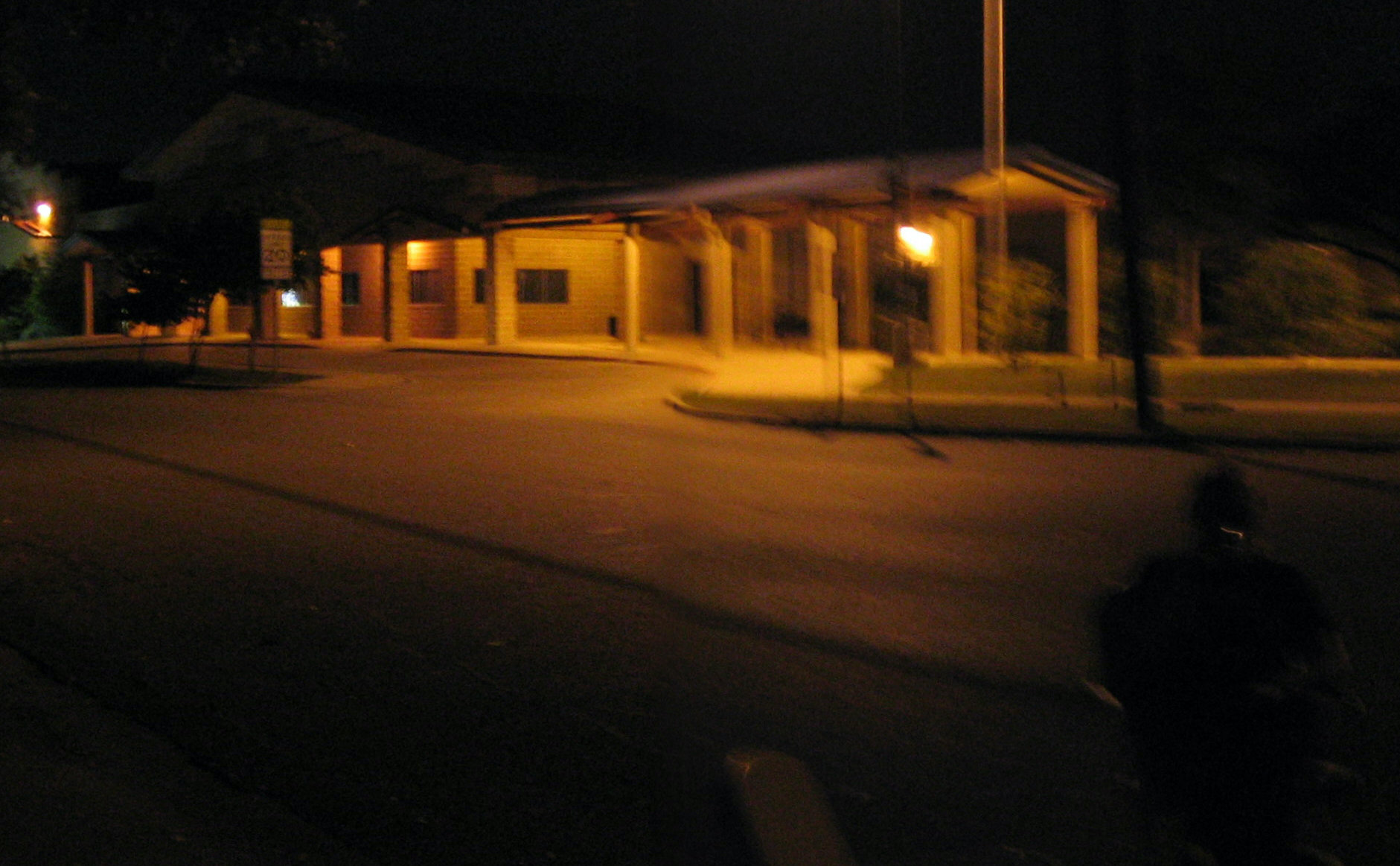 a blurry photo of an elementary school building at night, in a small town in central Texas. the darkness is illuminated with several sodium lights that cast an eerie but comforting glow onto the street in front of it. a person is just barely visible in silhouette in the corner of the frame.