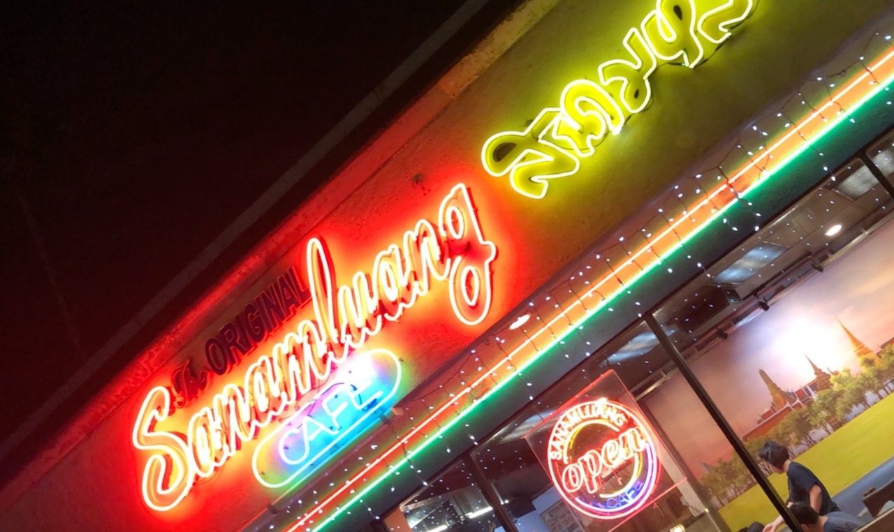 a cropped photo of Sanamluang Thai diner in north Hollywood. part of a neon sign is visible on the restaurant's facade. a circular open sign in neon hangs on a window.