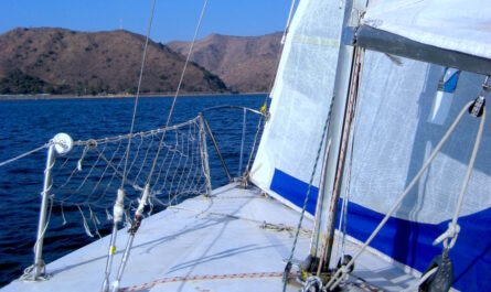 a photograph from the bow of a small sailing craft on calm blue waters. grassy brown hills dotted with green shrubs on the horizon separate the light blue sky and the sea below. a white and blue sail hangs from the mast, with rigging here and there.