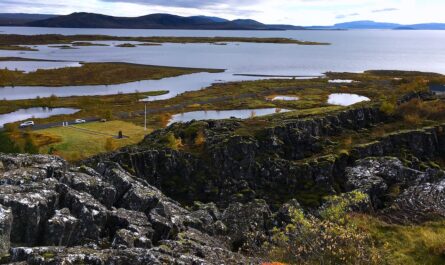 photo of the lava-strewn Thingvellir National Park in Iceland. mossy and jagged volcanic rock dominates the lower two-thirds of the image, with tiny vehicles in the distance for scale. beyond, the top part of the photo is where horizon, mountain, and water meet. here, the north american and eurasian tectonic plates are drifting apart.