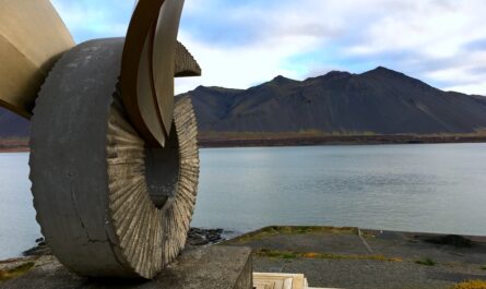 photograph of a sculpture in Borgarnes, Iceland titled "Brákin." the sculpture overlooks a body of water and then a mountain range covered in black dirt. the sculpture is a curl of concrete, six feet tall, curled with ridged sides to resemble a ram's horn. a piece of metal looks like stylized wings coming off the horn, which is meant to resemble a cloth being pulled away from the center of the horn. the sculpture itself is on a stone pedestal, which is itself on a wide wooden pedestal.