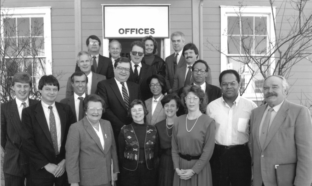 a group of nineteen people stand together in staggered rows outside a nondescript office. the men and women are all wearing some form of business attire. the photo is in black and white, but as far as i can tell it's from the nineteen-eighties.