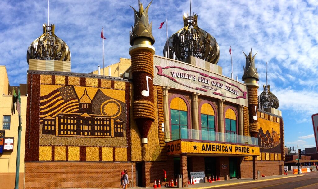 the world's only corn palace in Mitchell, SD. the building is ostentatious and tacky. red brick and thatched corn husk serve as most of the building's face. flanking the entrance are two friezes made of cobs of light and dark corn. atop the building are five metallic minarets. the sky is blue with light clouds stretching end to end. 