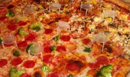 a pizza that may or may not look enormous, depending on the scale. the image is cropped close so only glimpses of the pizza box are visible along the edges. the toppings on half of the round pizza are pepperoni and broccoli. the toppings on the other half are pineapple, mushroom, and feta. if you look closely, you'd see that the pizza is cut into squares. it's also dotted with eight or so of the little plastic tables that keep a pizza lid from drooping onto your delicious pizza pie.