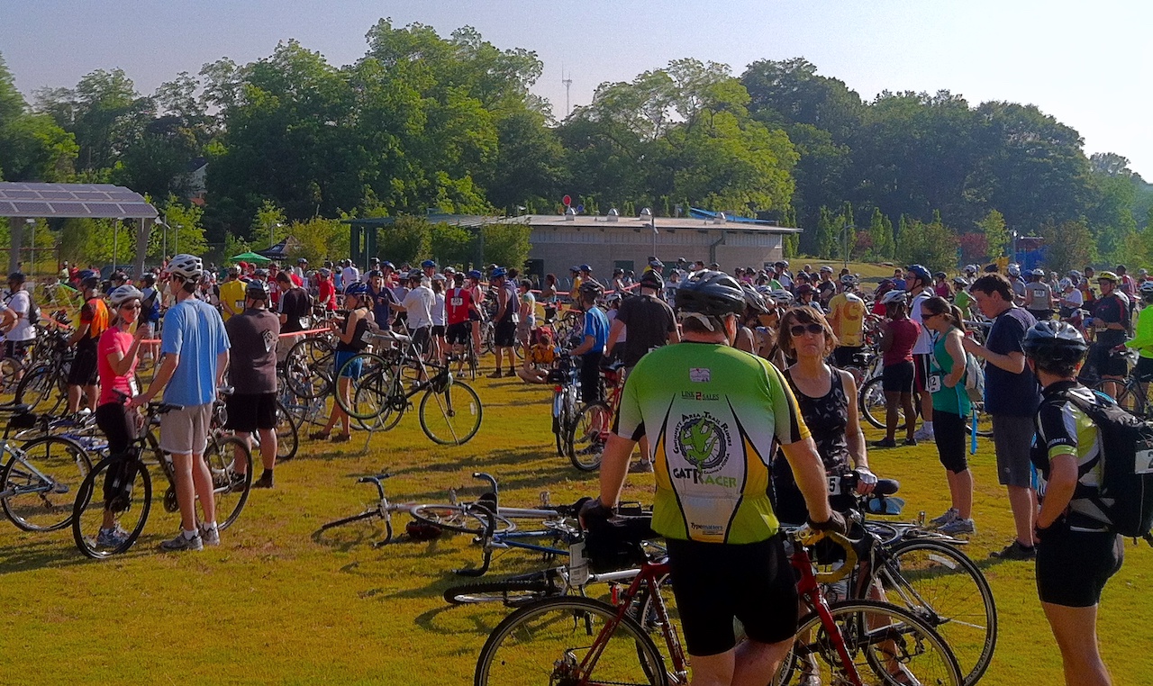 a crowd of cyclists gather in a flat green space edged with trees