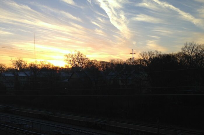 streaks of clouds in a blue and orange sky at dusk. train tracks are barely visible beneath the treeline.