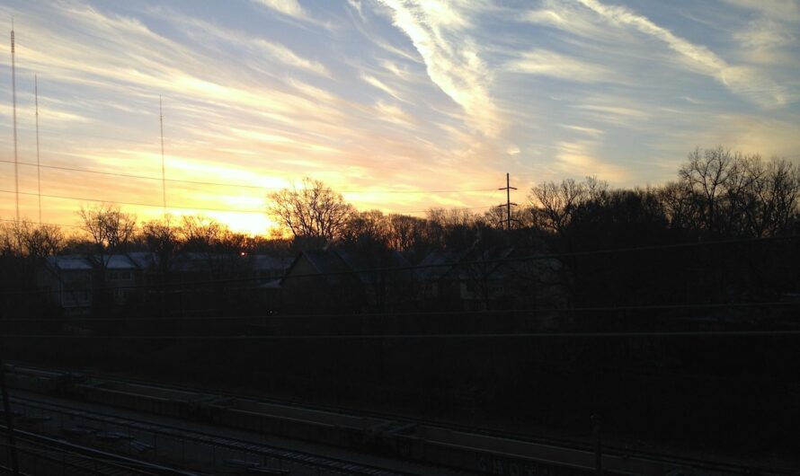 streaks of clouds in a blue and orange sky at dusk. train tracks are barely visible beneath the treeline.